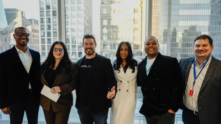 Group of six professionals posing for a photo in an office setting with large windows, showcasing a city skyline. They are dressed in business attire, smiling, and appear to be engaged in a networking event or conference.