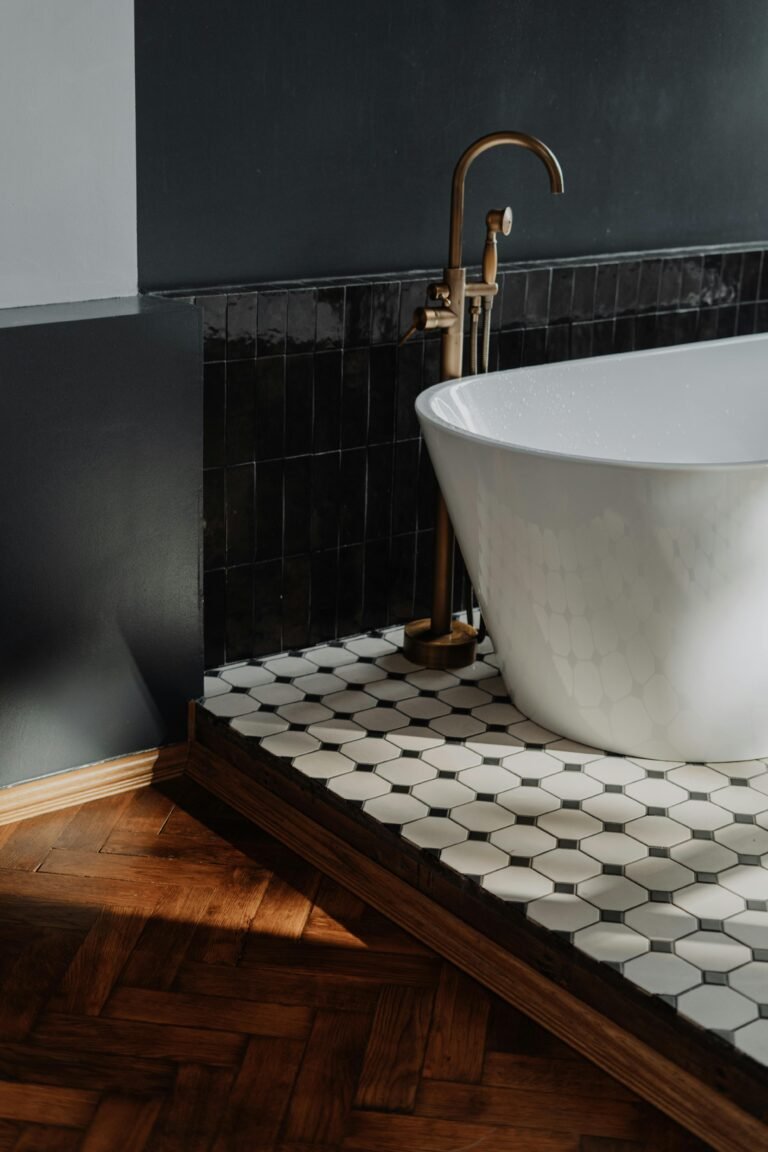 Modern bathroom featuring a freestanding white bathtub with a sleek design, complemented by a stylish gold faucet. The floor showcases a unique hexagonal tile pattern in black and white, set against dark walls and warm wooden accents, creating a sophisticated and inviting atmosphere.