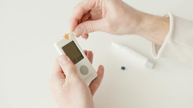 Person using a glucose meter to check blood sugar levels, holding the device in one hand while preparing a test strip with the other, with a pen-like injector and a blue tablet visible in the background.