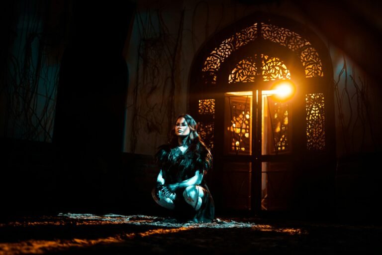 A woman in a dark, feathered outfit squats in a dimly lit room, illuminated by warm light from an intricately designed doorway. The scene captures an atmospheric, mysterious vibe with shadows and textured walls, enhancing the dramatic effect.