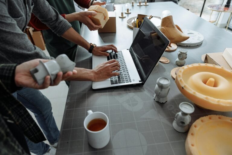 Team members collaborating on a design project at a workspace, using a laptop while handling various prototype models and materials, with a mug of tea visible on the table.