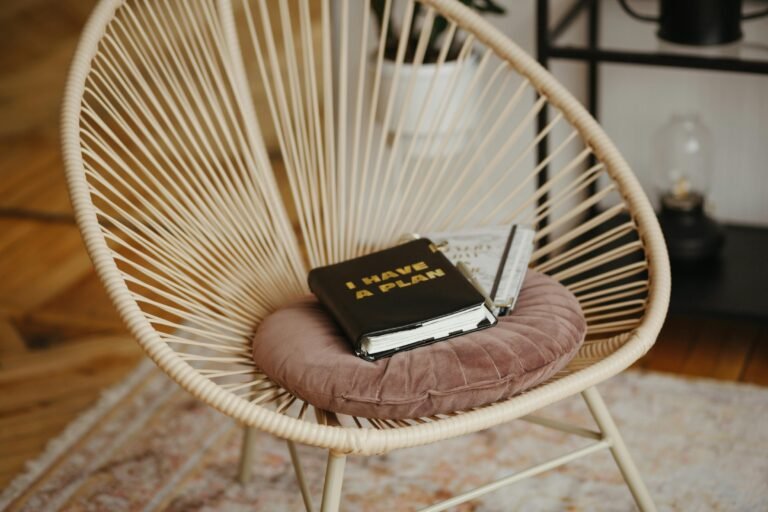 Stylish rattan chair with a plush pink cushion, featuring a black planner titled "I HAVE A PLAN" and a notebook resting on it, set in a cozy, modern interior.