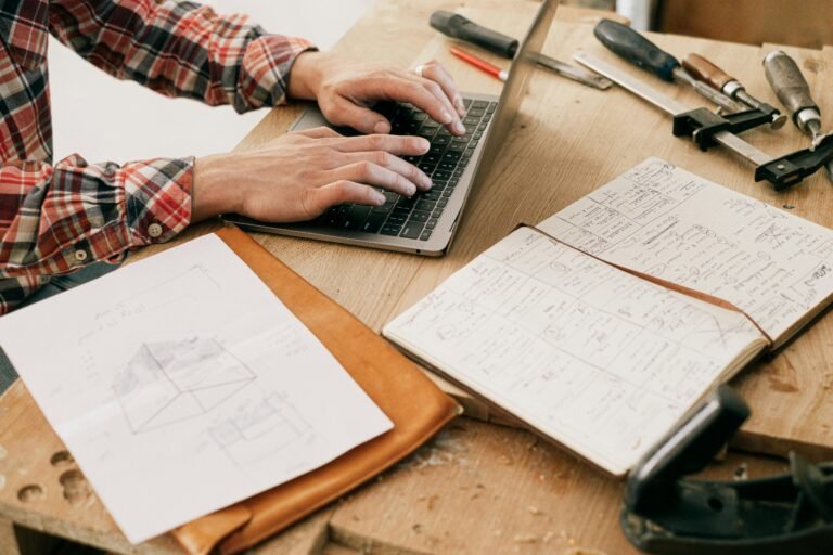 A person in a plaid shirt types on a laptop, surrounded by woodworking tools, a sketch of a design, and an open notebook filled with notes and plans.