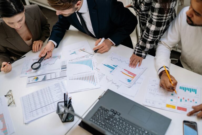 Business professionals analyzing financial data and charts on paper in a collaborative meeting. Various graphs and reports are spread across a table, with a laptop and office supplies visible, highlighting teamwork and data-driven decision-making.