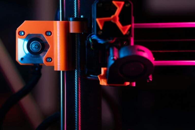 Close-up of a 3D printer's extruder and stepper motor, featuring vibrant orange components against a dark background with colorful lighting. The image highlights the intricate design and technology behind 3D printing mechanisms.