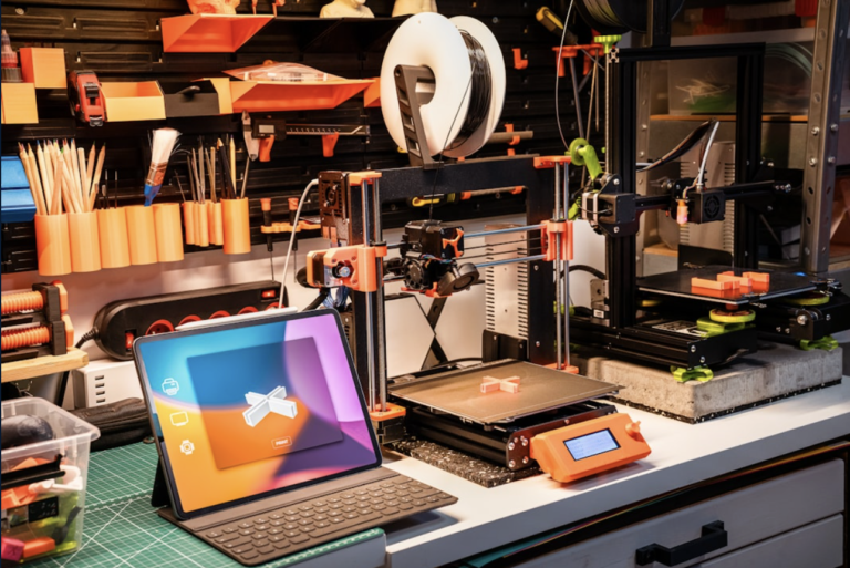 A well-organized 3D printing workspace featuring two 3D printers, a tablet displaying a design interface, and various tools and materials on a workbench. The setup includes colorful storage solutions and a green cutting mat, ideal for makers and hobbyists.