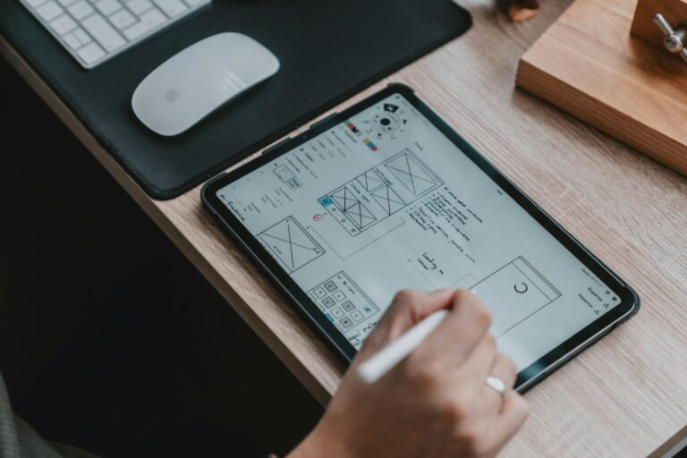 Person sketching wireframes on a tablet using a stylus, with a keyboard and mouse in the background, illustrating the design process in a modern workspace.